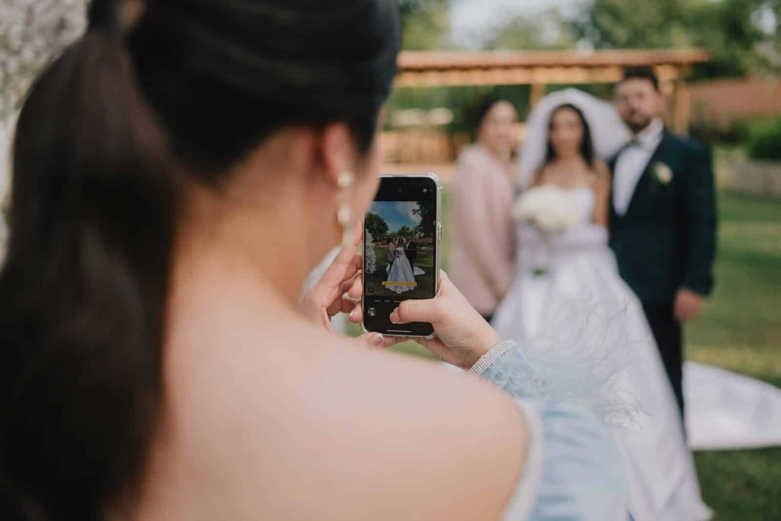 Gast fotografiert das Paar Gast fotografiert Braut und Bräutigam mit dem Handy vor einer Pergola